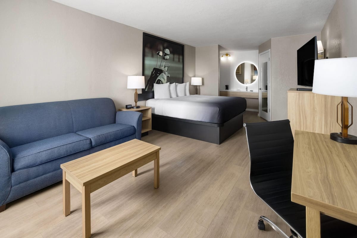 A modern hotel room with a blue sofa, a wooden coffee table, a bed with a black headboard, a desk with a lamp, and a TV.