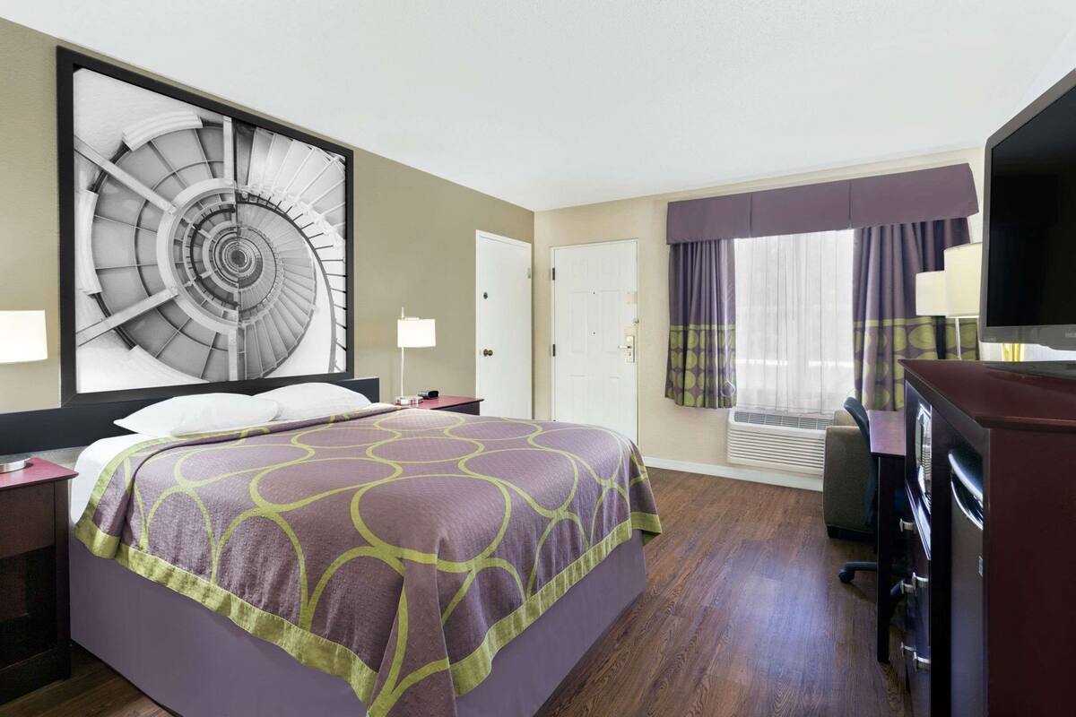 A hotel room with a large bed, a desk, a television, and a large window with curtains.