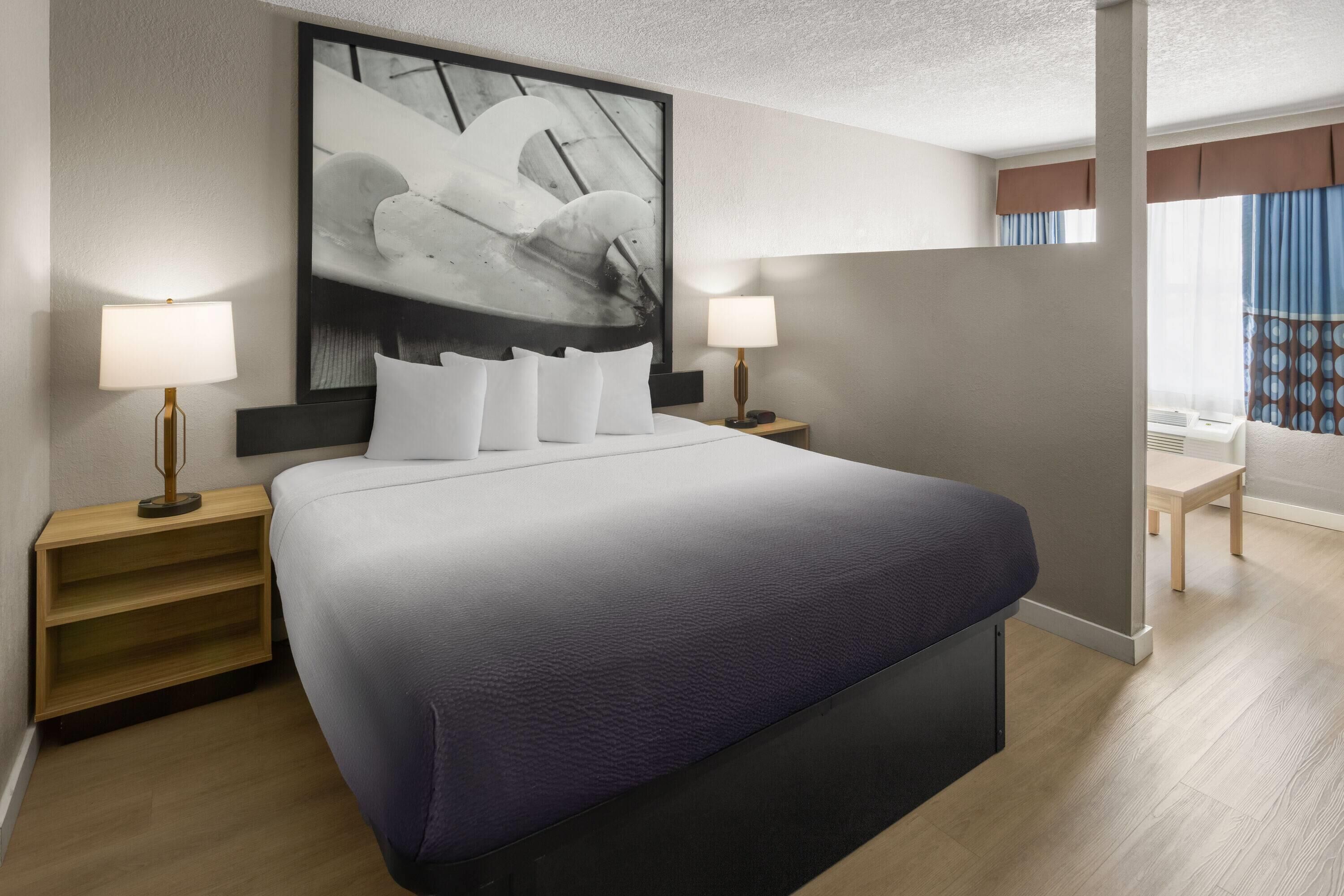 A modern hotel room with a large bed, two bedside lamps, a wall-mounted artwork, and a view of the outdoors.