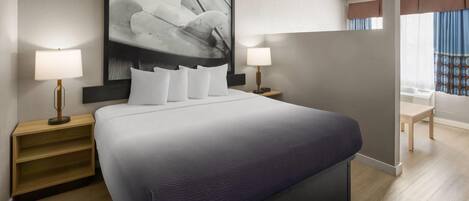 A modern hotel room with a large bed, two bedside lamps, a wall-mounted artwork, and a view of the outdoors.