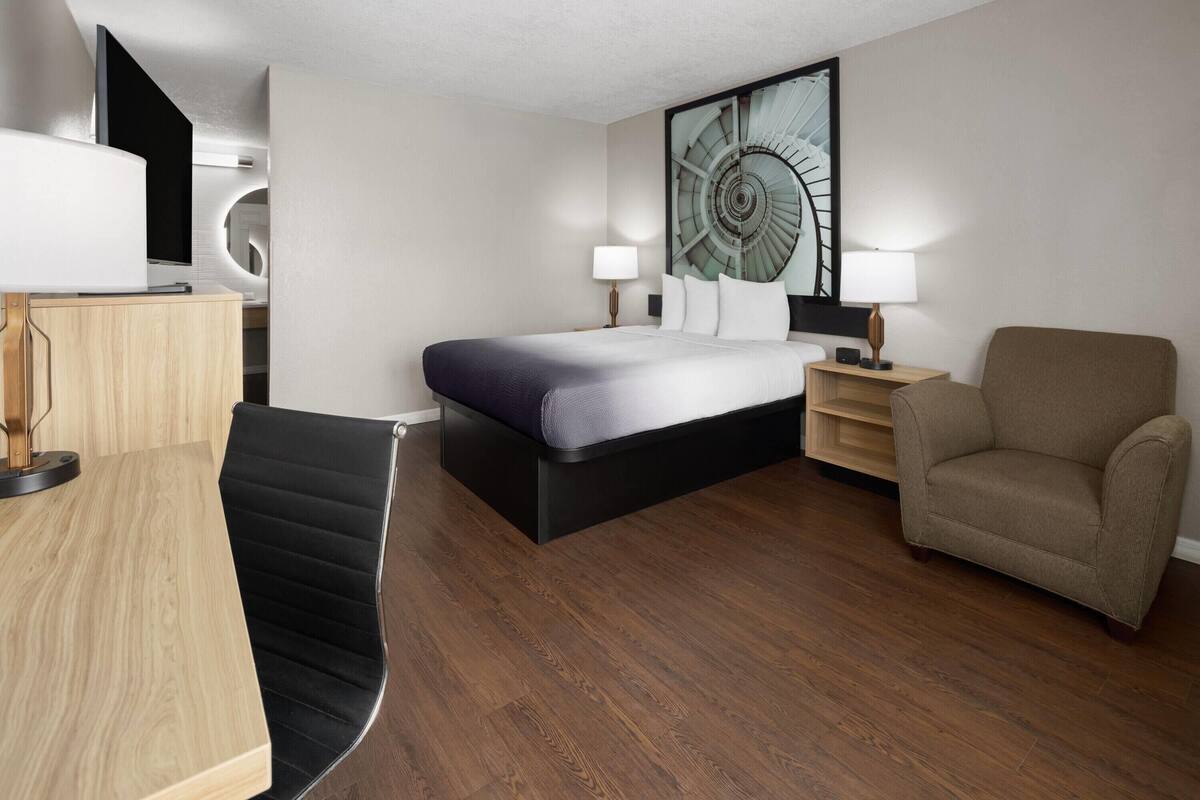 A modern hotel room with a bed, a desk, a chair, a sofa, and a large wall art piece.