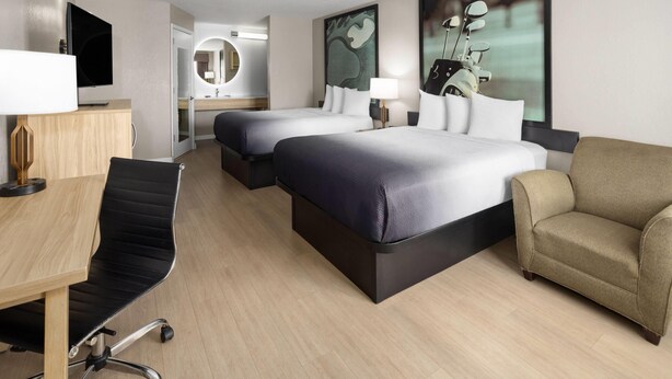 Habitación de hotel con dos camas, un escritorio, una silla, un televisor, una lámpara y obras de arte en las paredes.