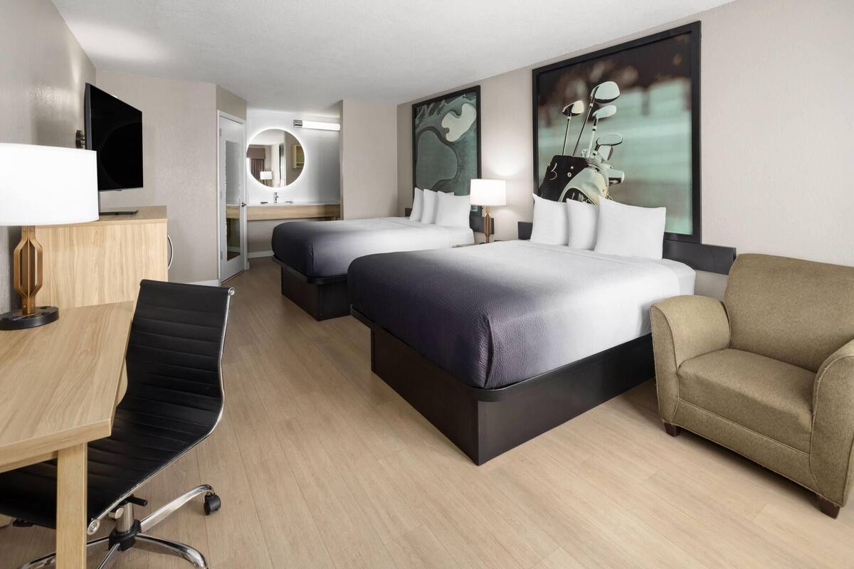A hotel room with two beds, a desk, a chair, a TV, a lamp, and artwork on the walls.