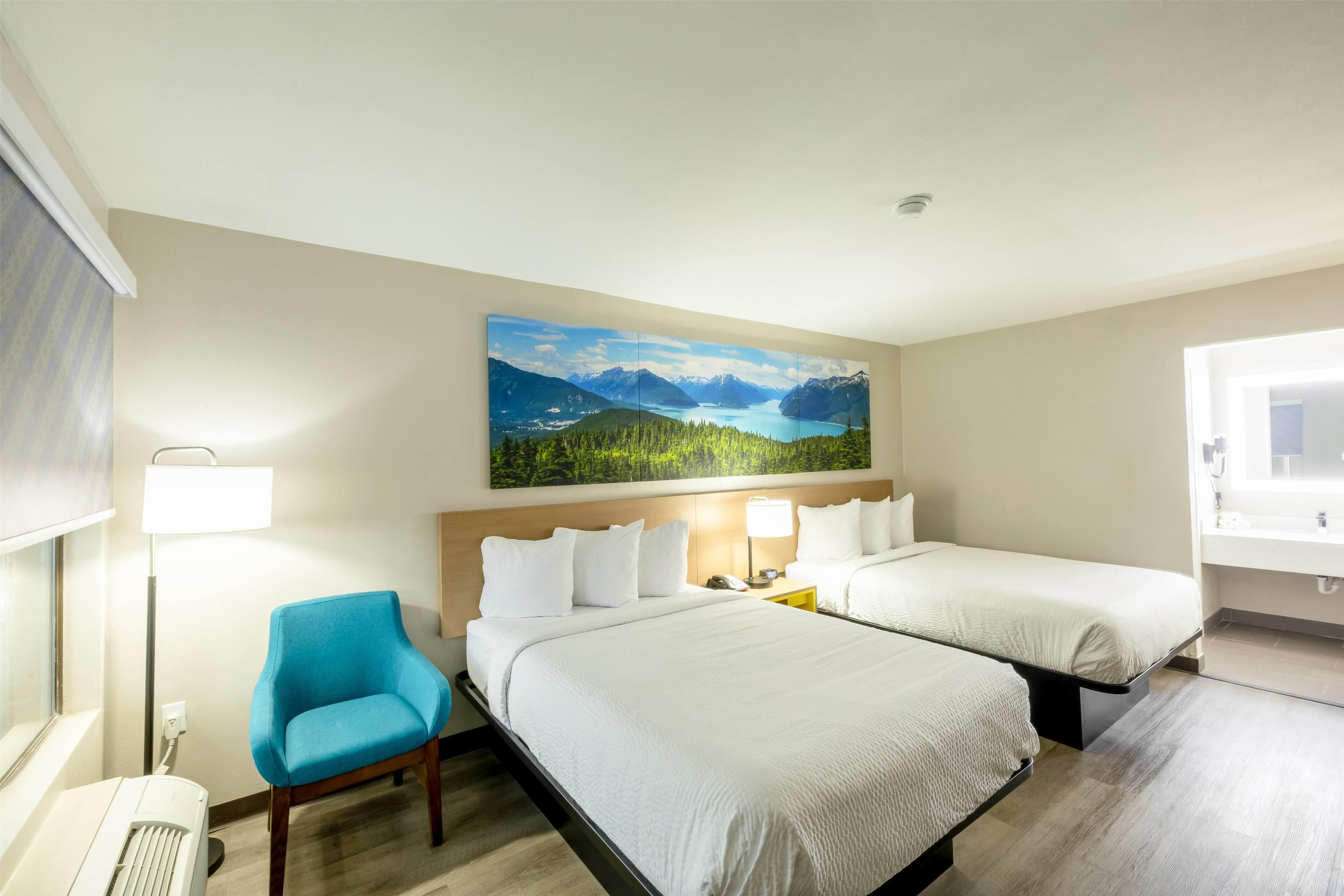 A hotel room with two beds, a blue chair, a painting of a mountain landscape, and a window with a view of the room.