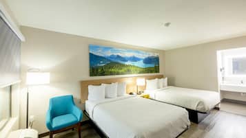 A hotel room with two beds, a blue chair, a painting of a mountain landscape, and a window with a view of the room.