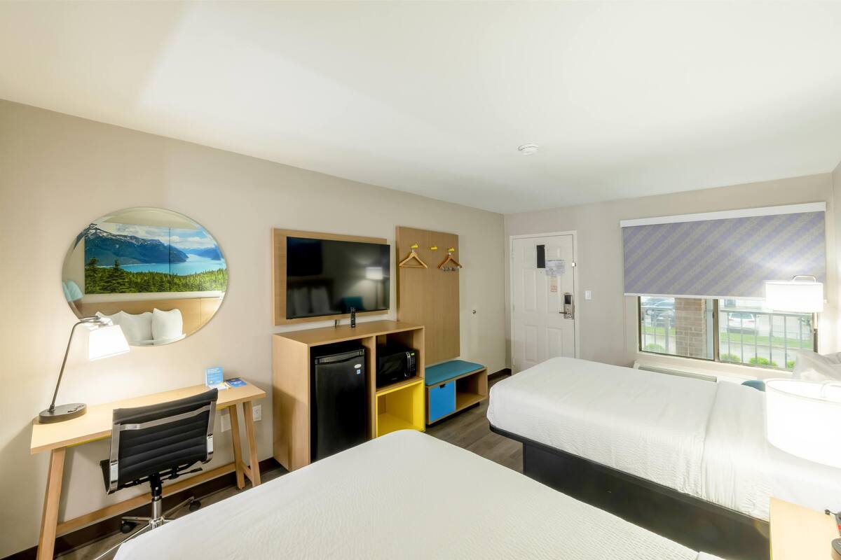 Ein Hotelzimmer mit zwei Betten, einem Schreibtisch mit Computer, einem Fernseher, einem Spiegel und einem Fenster mit Ausblick.