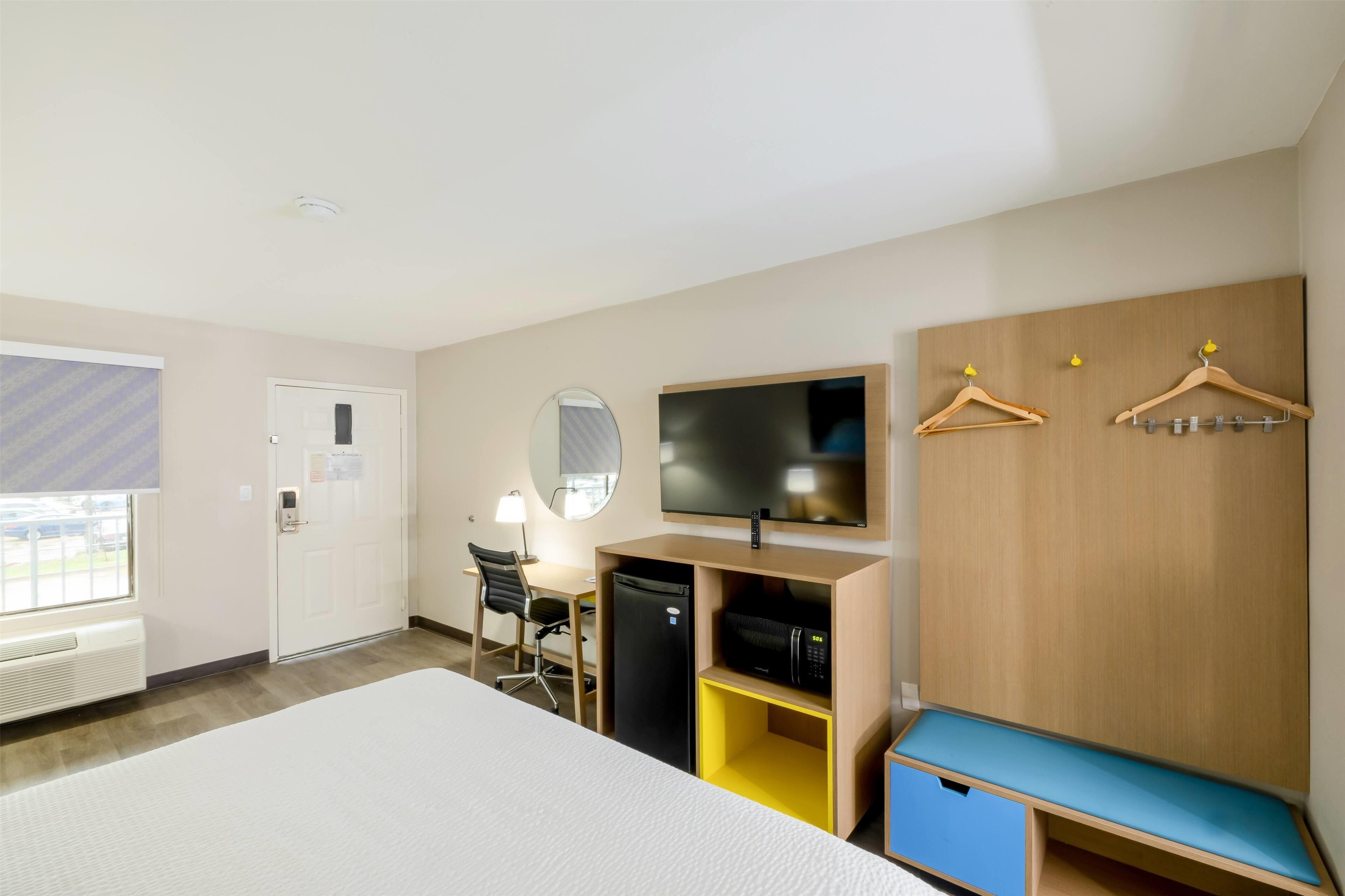 A hotel room with a bed, a desk with a chair, a TV, a wardrobe, and a seating area with a bench.