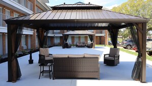 Eine Terrasse im Freien mit überdachter Sitzgruppe, inklusive einer Couch und Stühlen, und ein Gebäude mit Ziegelverkleidung im Hintergrund.