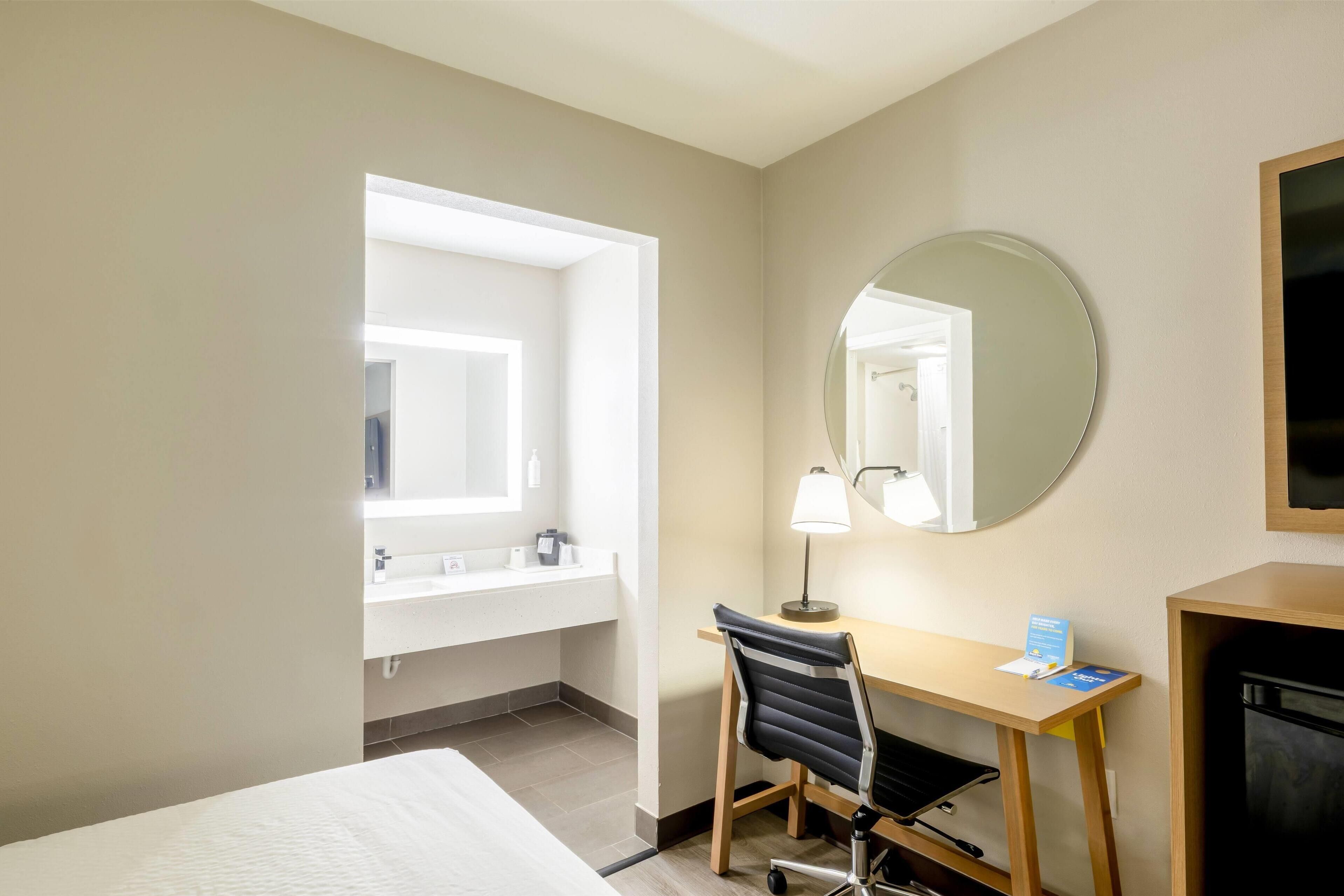 A hotel room with a bed, a desk with a chair, a round mirror, a sink, and a television.