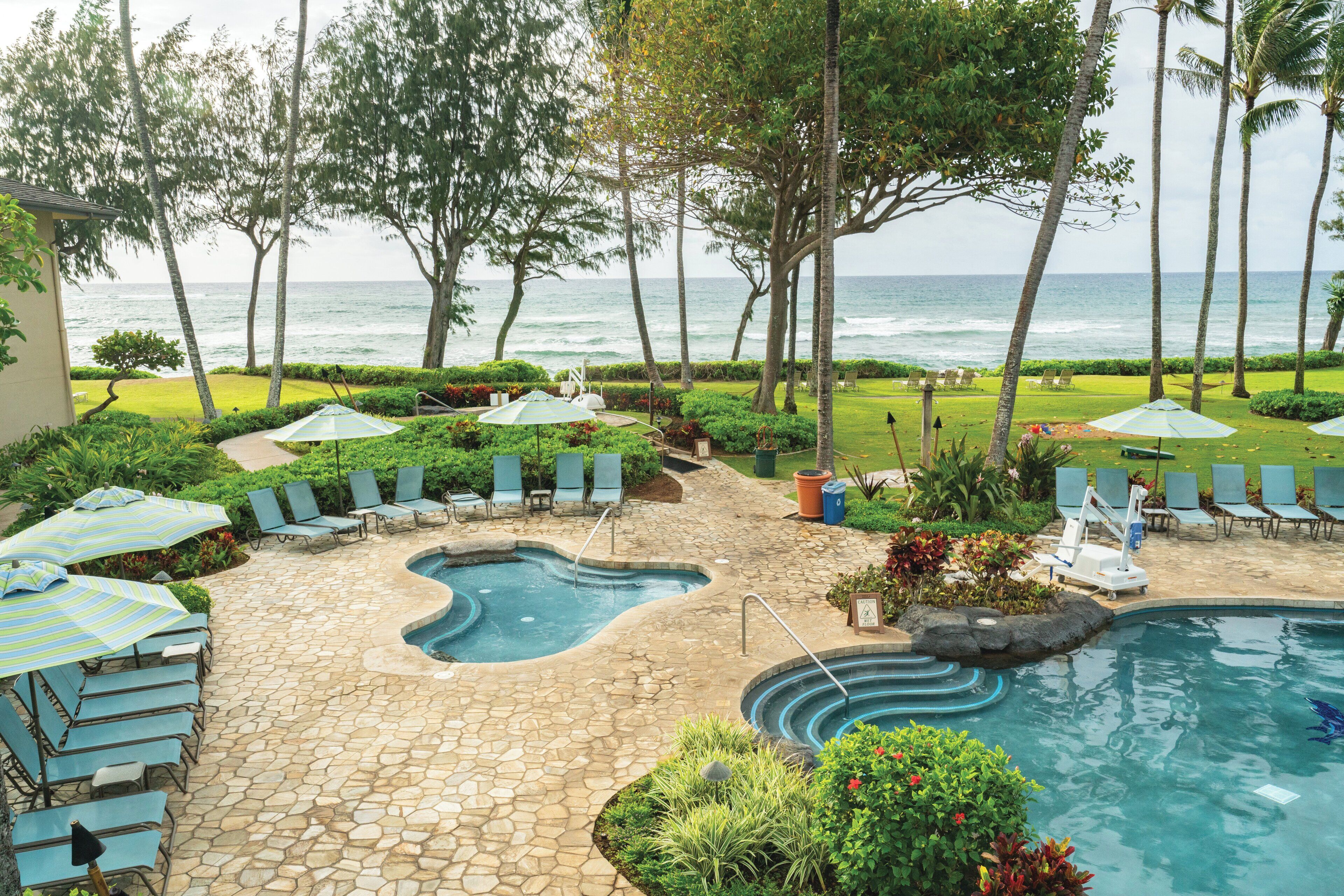 A pool area with lounge chairs and umbrellas, surrounded by palm trees and a view of the ocean.