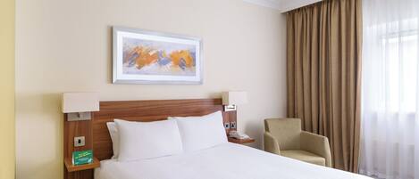 A hotel room with a large bed, a headboard, a framed abstract painting, a chair, a nightstand, and a lamp.