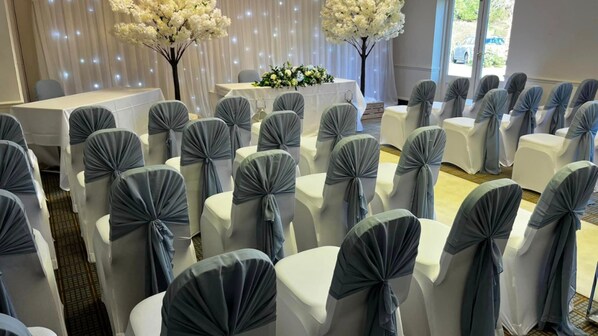 A conference room with rows of white chairs with grey sashes, a white table with a floral centerpiece, and a backdrop of white drapes with string lights. - Holiday Inn Colchester by IHG (Colchester)