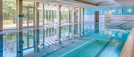 A large indoor swimming pool with clear glass walls, a diving board, and a sunlit area with lounge chairs.