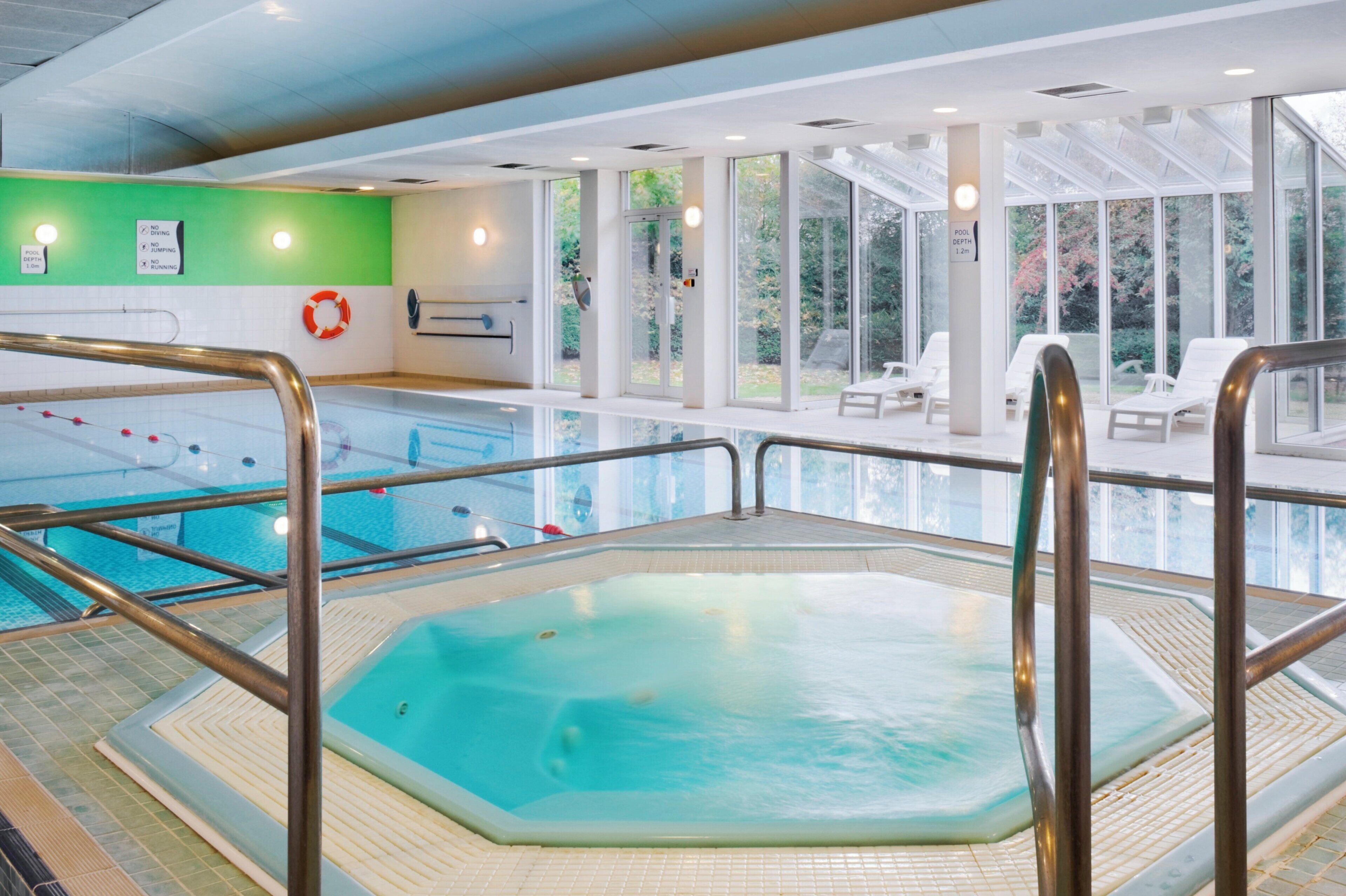 Una piscina interior con un jacuzzi, señales de seguridad y una zona soleada con sillones de descanso.
