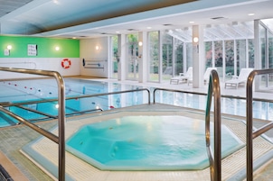 Una piscina interior con un jacuzzi, señales de seguridad y una zona soleada con sillones de descanso.