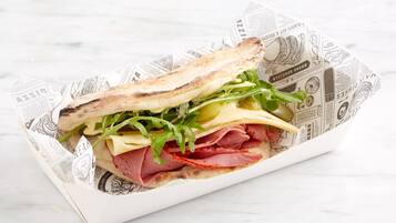 A sandwich with various ingredients, including cheese, greens, and meat, placed on a surface with newspaper-like material.