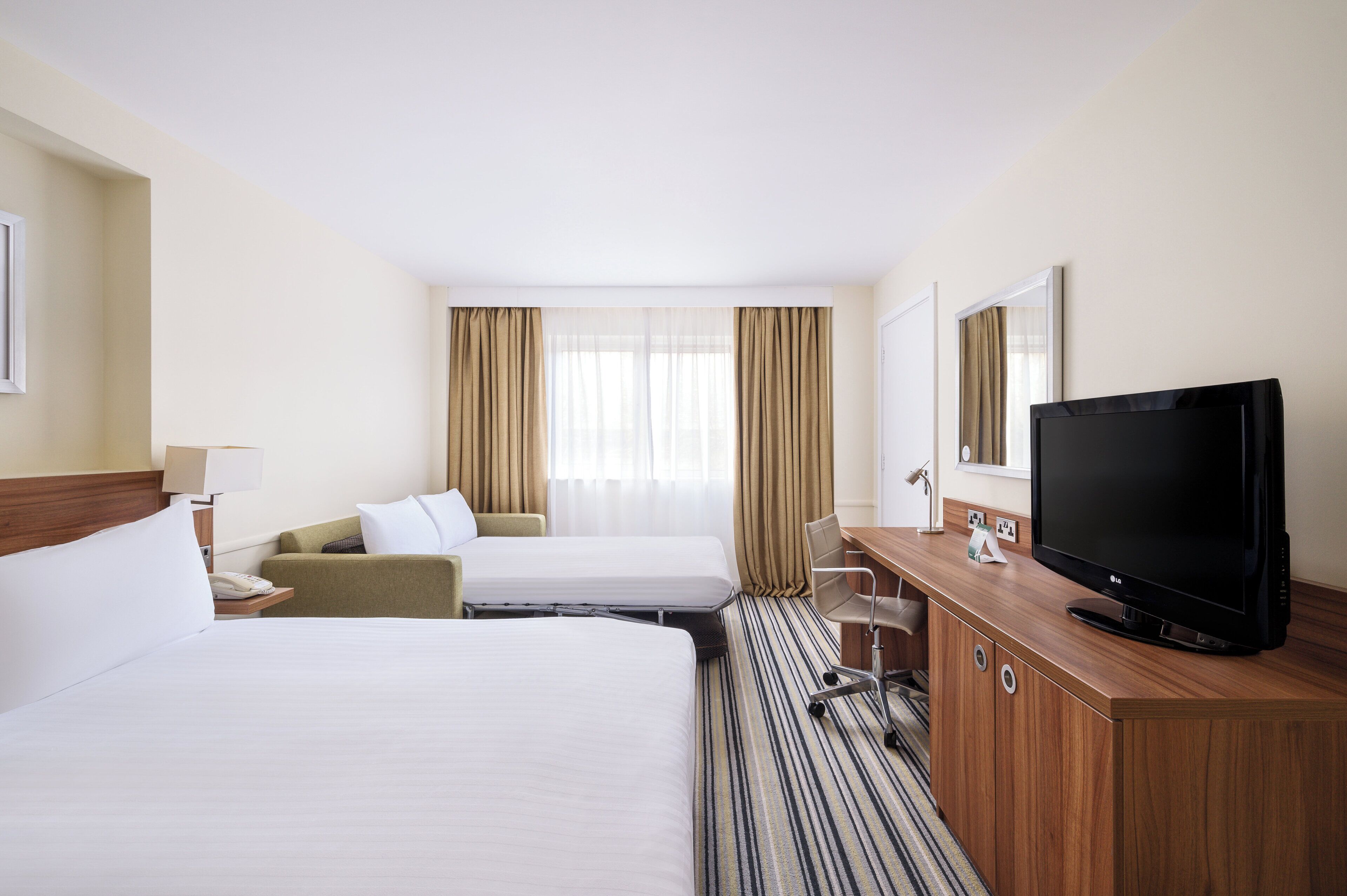 A hotel room with two beds, a desk with a television, a chair, and a window with curtains.