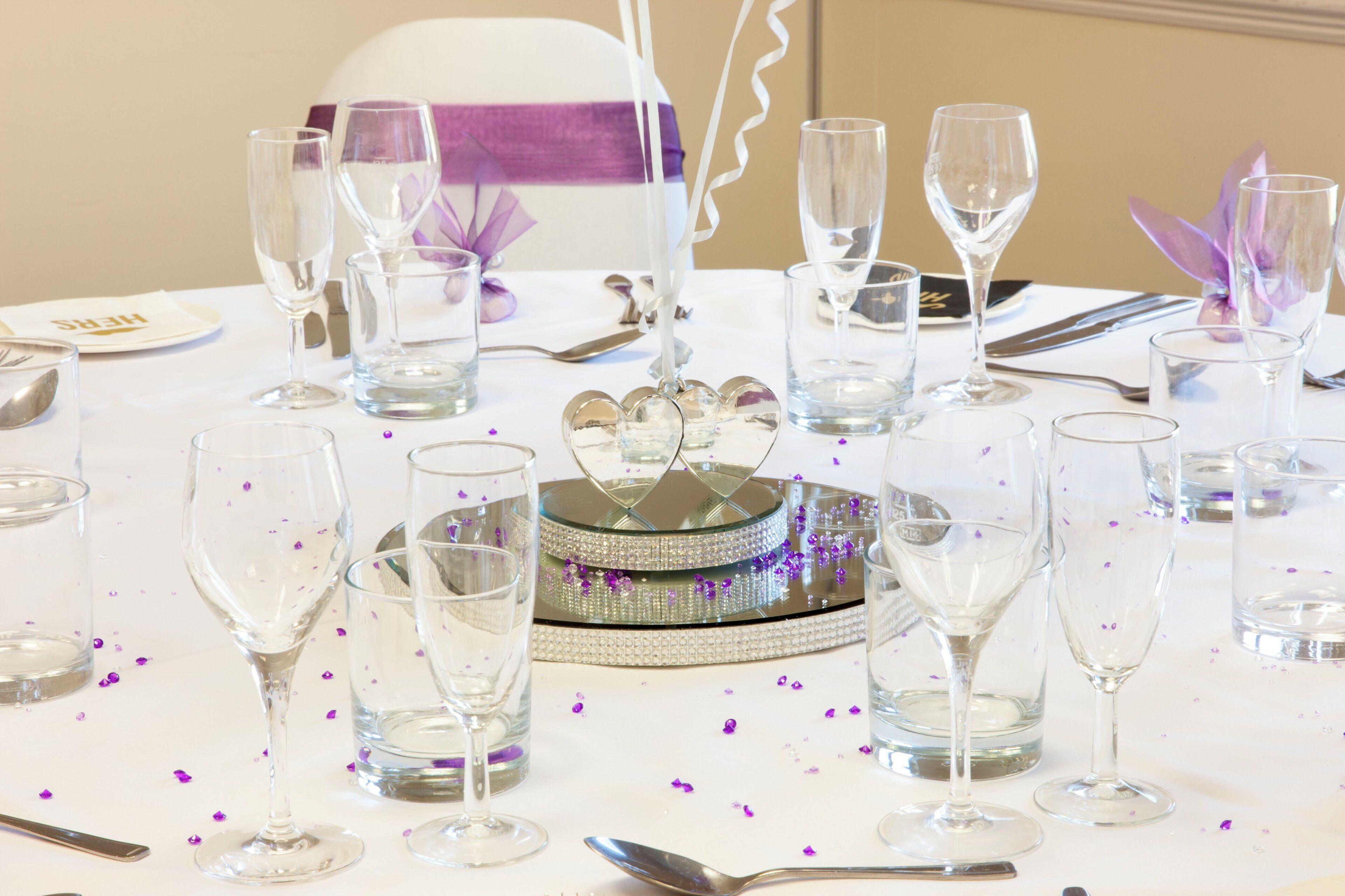 Tavola imbandita per un evento formale, con bicchieri trasparenti, un elemento decorativo al centro e una tovaglia bianca.