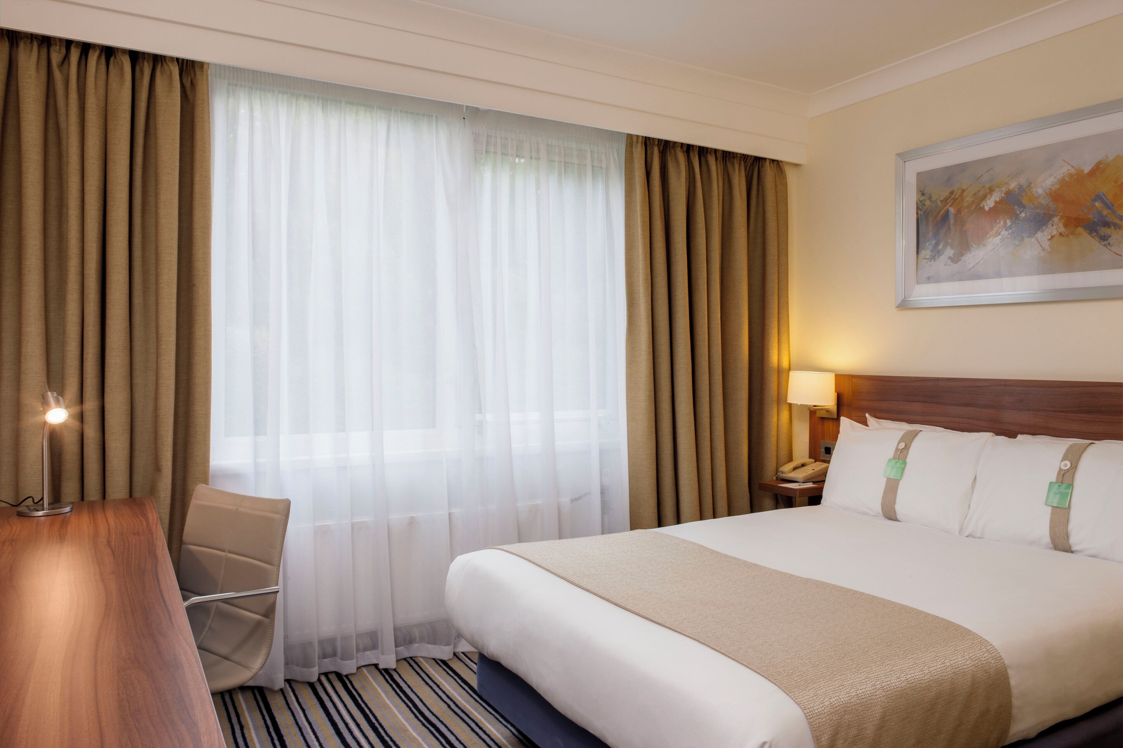 Habitación de hotel con cama, escritorio, silla, mesita de noche con lámpara y un cuadro en la pared.
