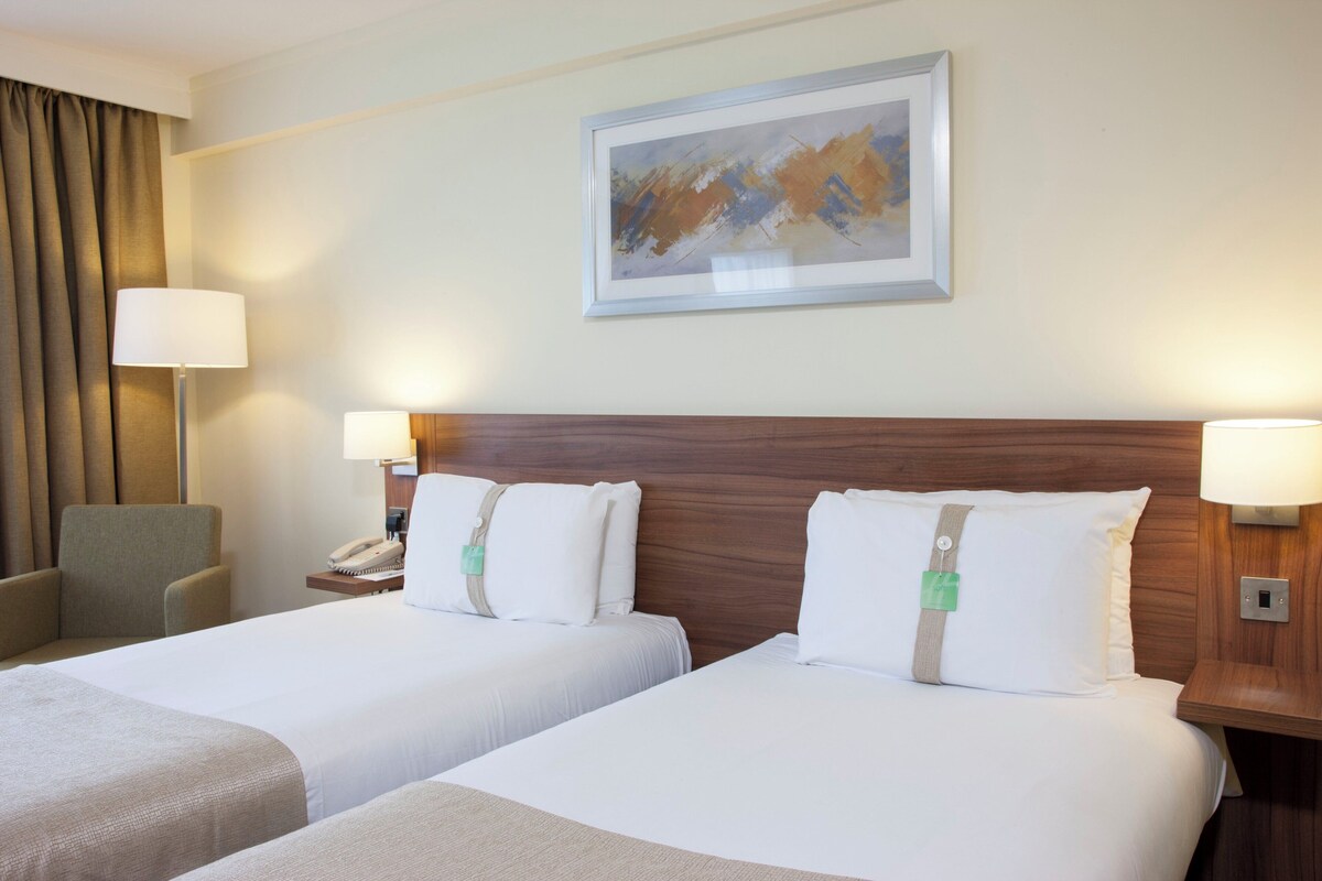 A hotel room with two beds, a wooden headboard, a painting on the wall, and a chair.