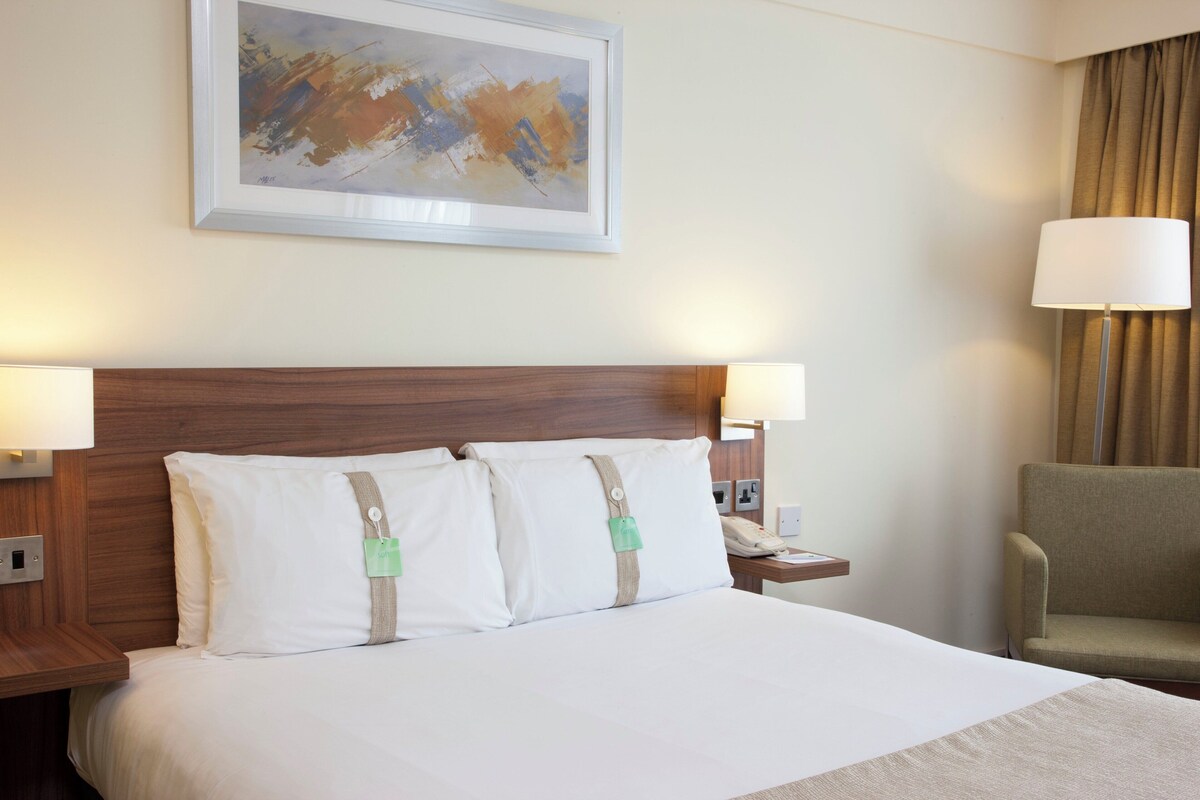 A hotel room with a large bed, two bedside lamps, a nightstand with a phone, a chair, and a painting on the wall.
