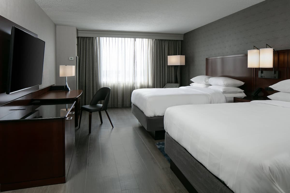 A hotel room with a flat-screen TV, a desk, two beds, a chair, and a large window with curtains.