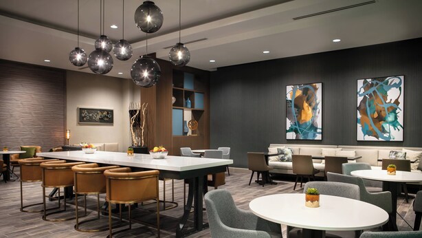 A modern hotel lobby with a long dining table, bar stools, round tables, and comfortable seating. - West Des Moines Marriott (West Des Moines)