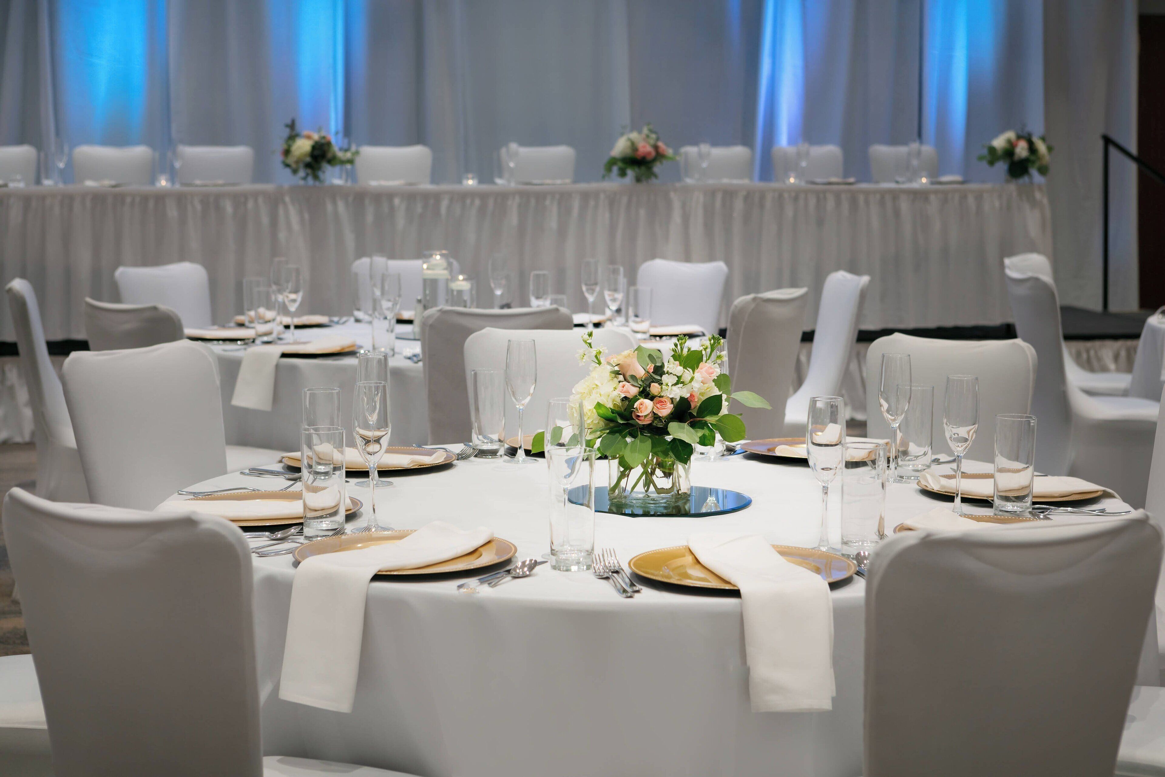 A banquet hall with round tables set for a formal event, complete with white tablecloths, gold cutlery, and floral centerpieces.