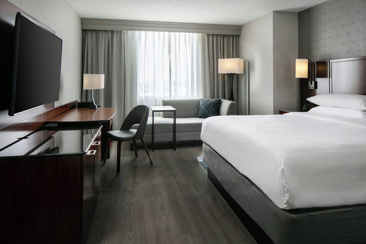 A hotel room with a large bed, a desk with a flat-screen TV, a chair, a small table, and two bedside lamps.