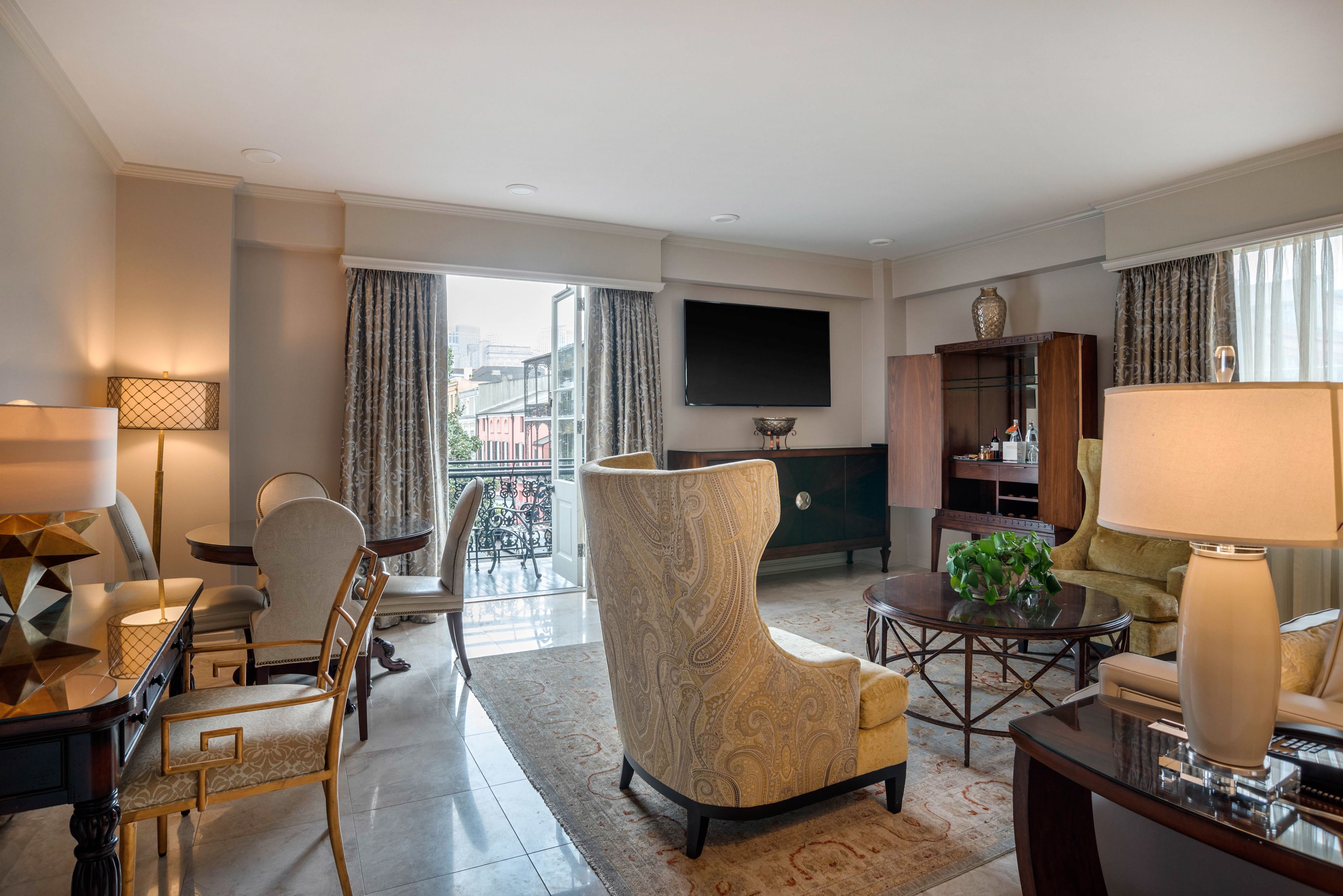 A modern hotel living room with a large TV, comfortable seating, a dining area, and a view of the city.