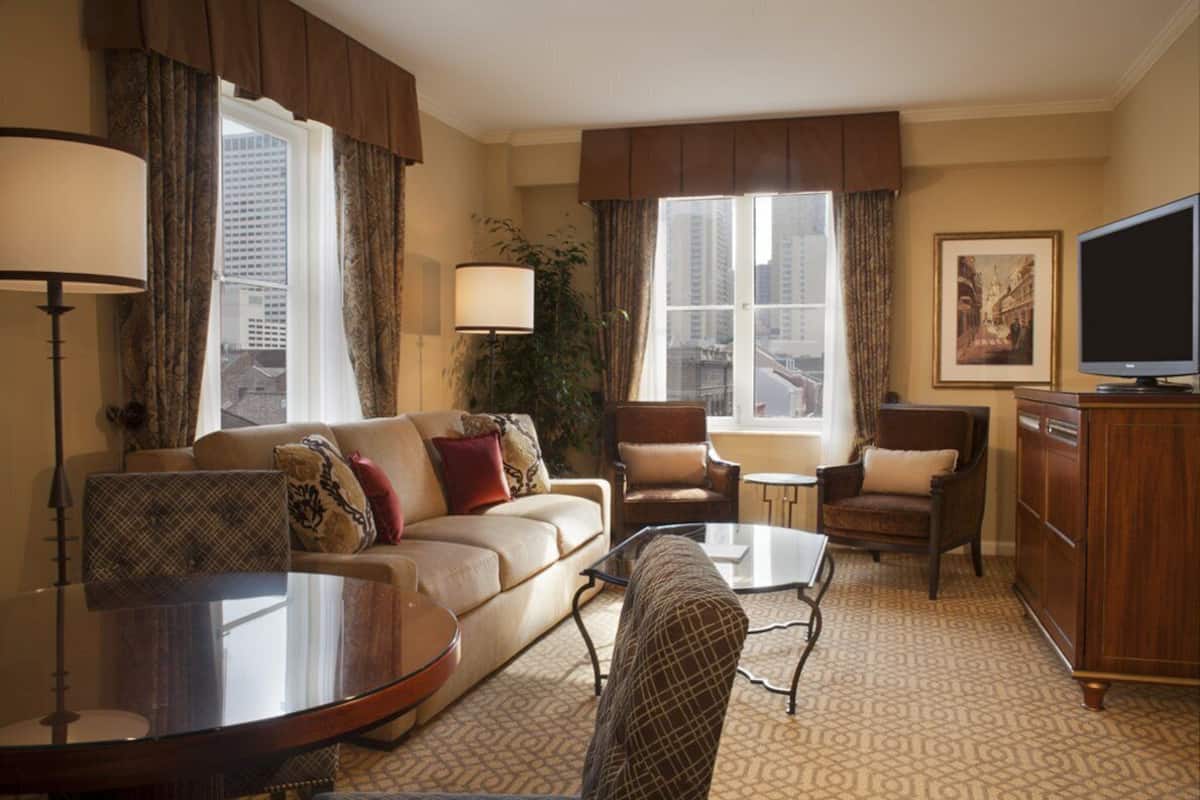 Habitació d'hotel amb sofà, butaques, taula de cafè, televisió i una finestra gran amb cortines.