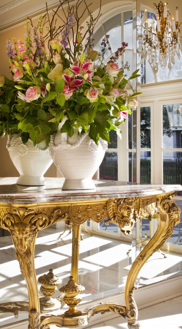 Una estancia imponente con muebles dorados ornamentados, grandes jarrones blancos con flores y un amplio ventanal con vistas al exterior.