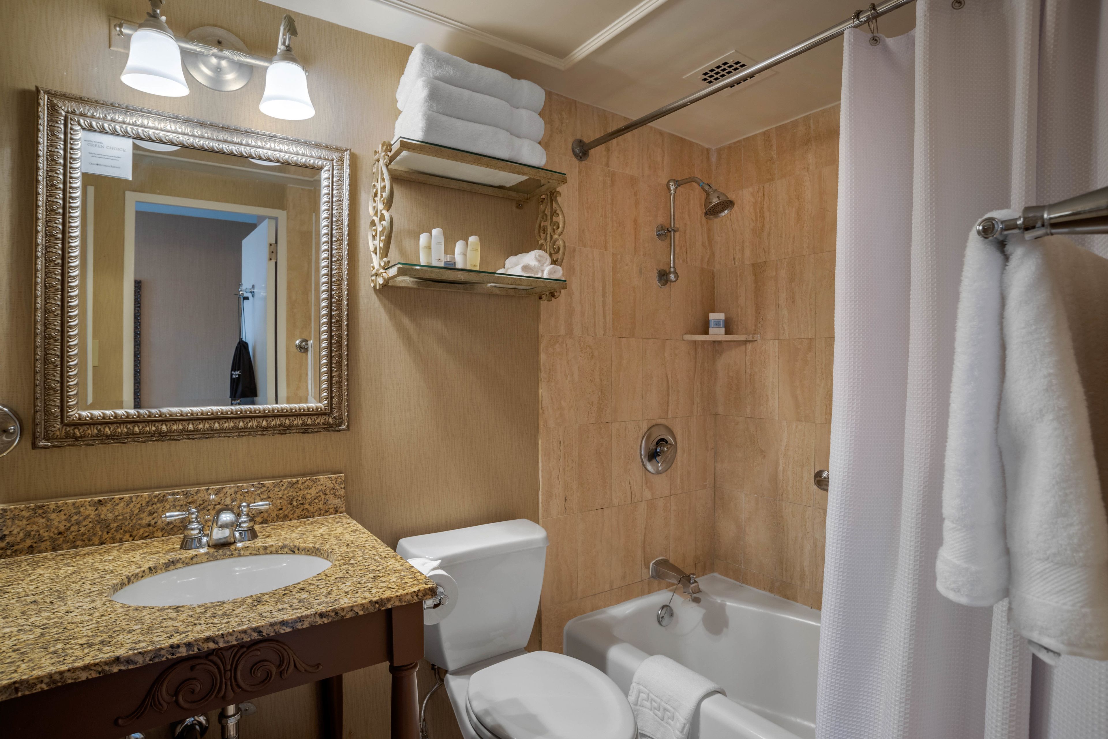 Un bany amb un lavabo amb taulell de granit, un vàter blanc, una banyera i una dutxa. Hi ha prestatges sobre el vàter i un suport de tovalloles al costat de la banyera.