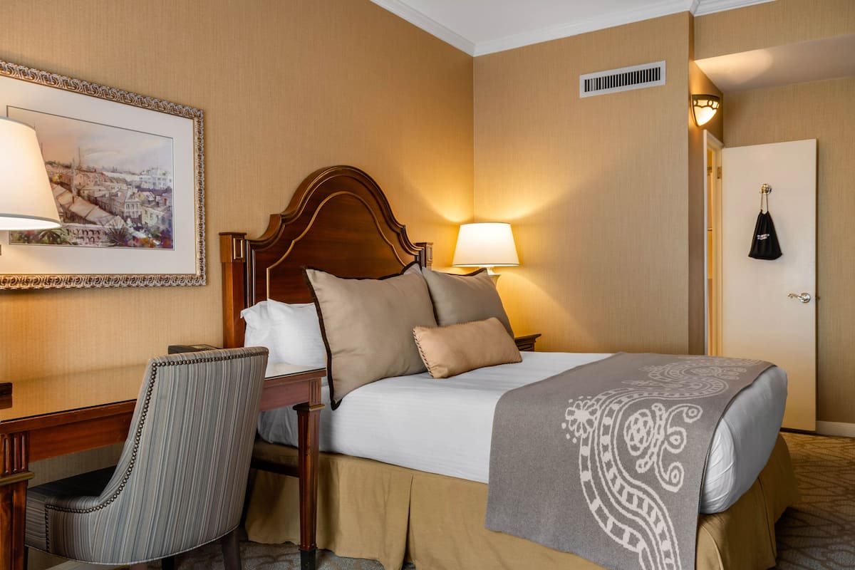 Habitació d'hotel amb un llit, dues làmpades de nit, una cadira, un quadre amb marc a la paret i una porta amb un abric penjat.