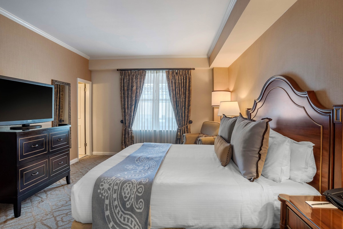 Une chambre d’hôtel avec un grand lit, un téléviseur à écran plat, une commode et une fenêtre avec des rideaux.