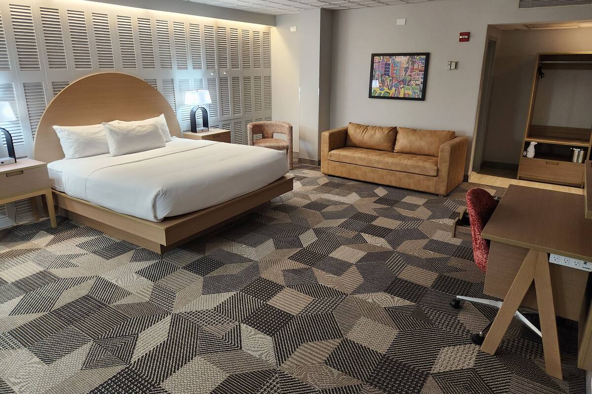 Quarto de hotel com uma cama grande, um sofá, uma escrivaninha e um tapete estampado.