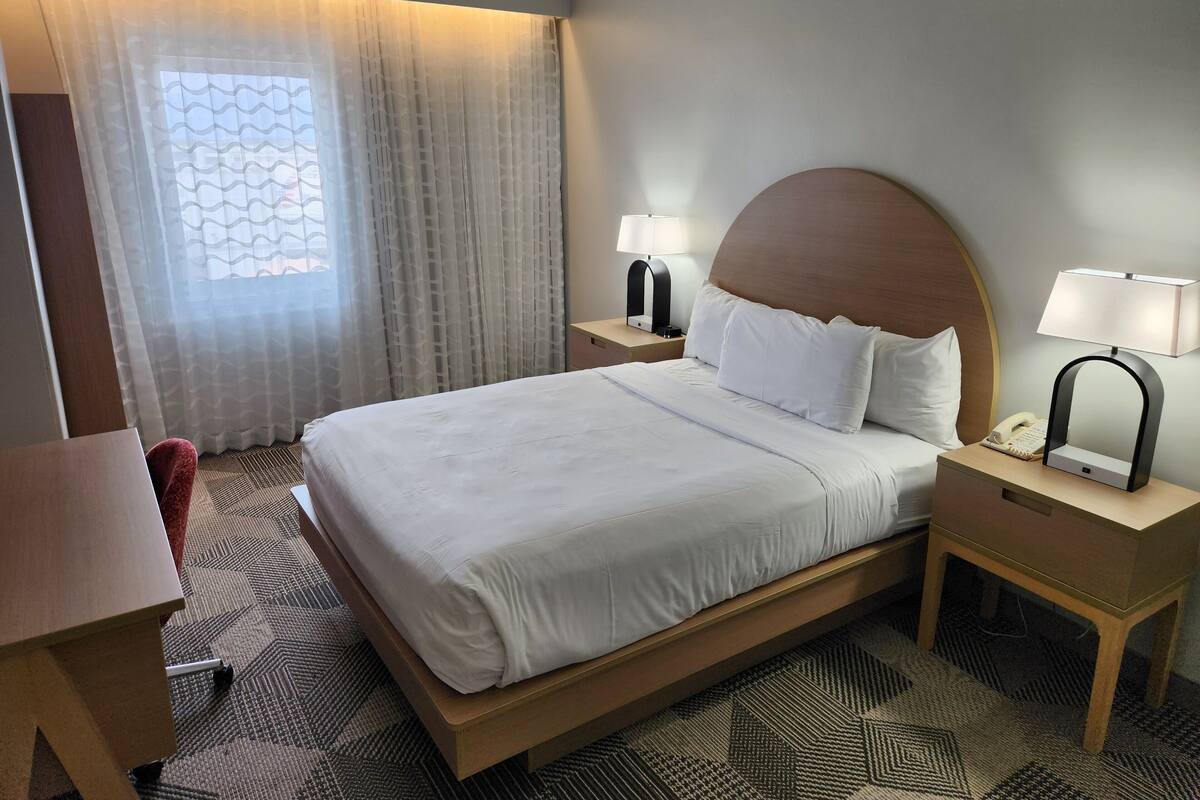 Quarto de hotel com uma cama grande, duas mesas de cabeceira com luminárias, uma escrivaninha e uma cadeira.