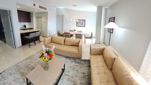 Una sala de estar moderna con un sofá beige, una mesa de centro con un centro de mesa floral y un comedor al fondo.