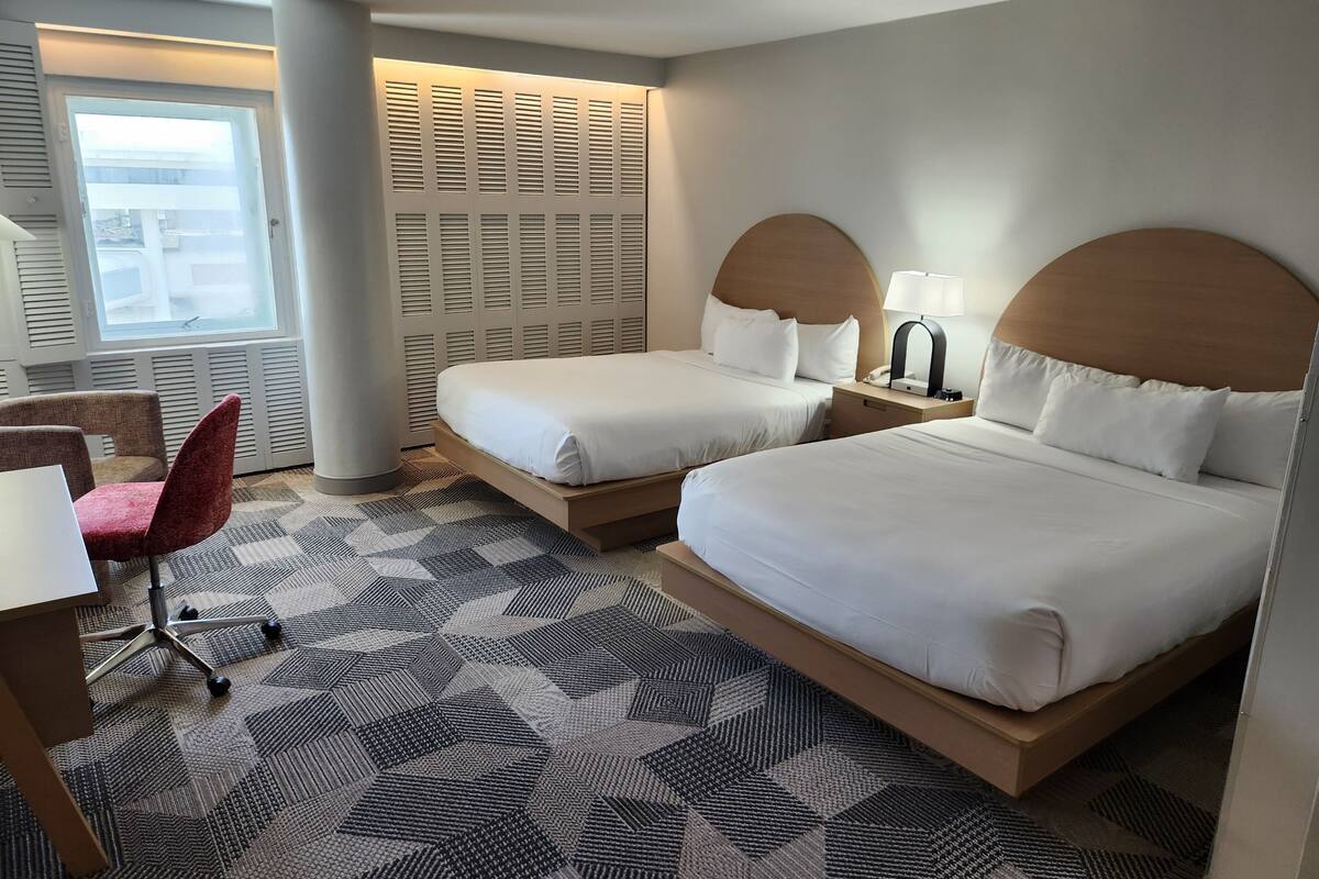 Quarto de hotel com duas camas, uma escrivaninha, uma cadeira e um tapete estampado.