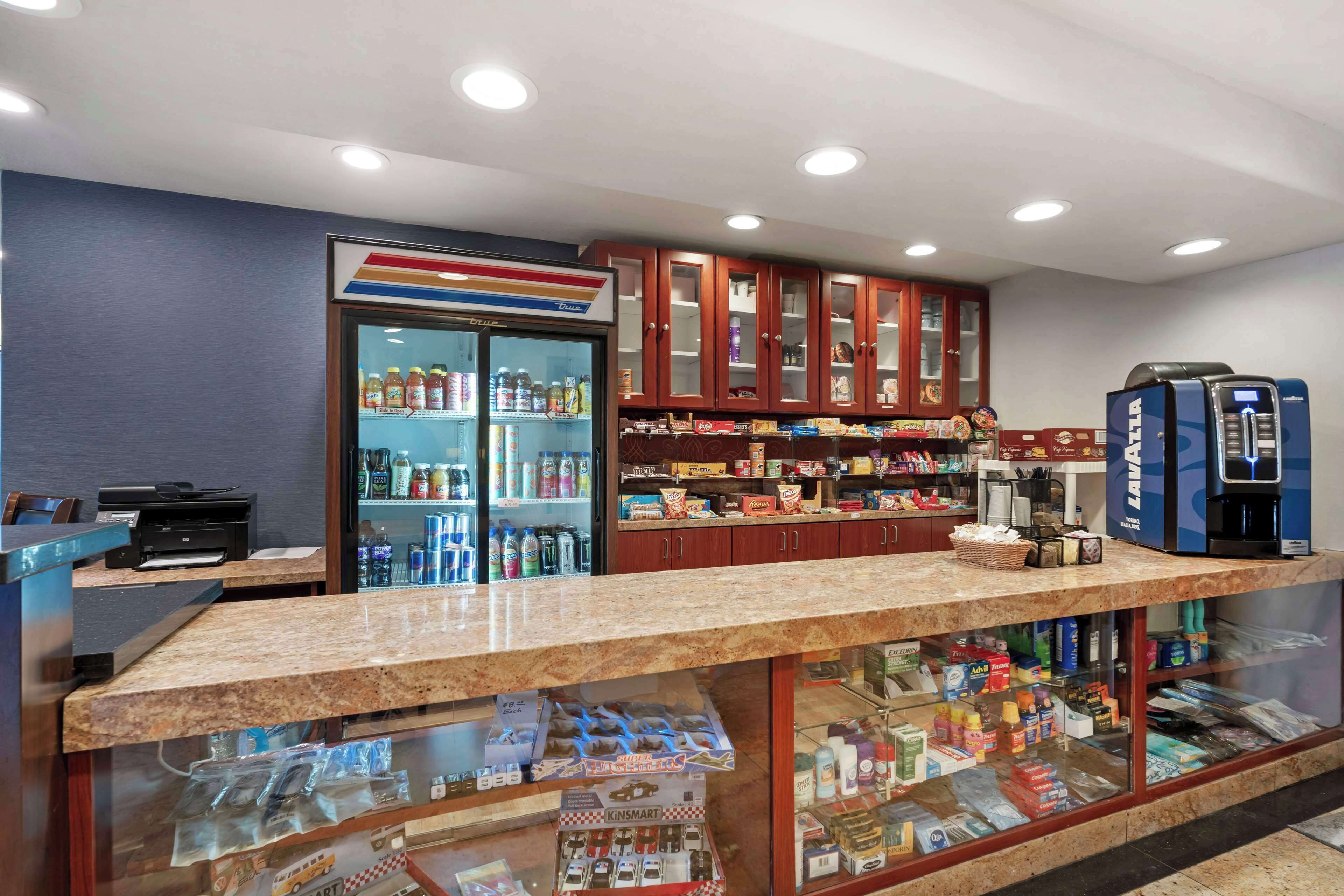 Zona de recepción con mostrador, refrigerador lleno de bebidas, máquina de café y varios snacks y artículos de aseo en estantes.