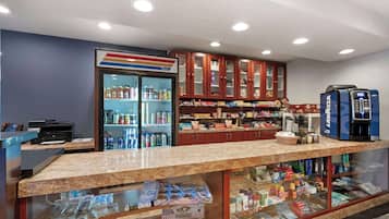 Zona de recepción con mostrador, refrigerador lleno de bebidas, máquina de café y varios snacks y artículos de aseo en estantes.