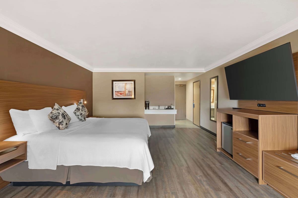 Une chambre d’hôtel moderne avec un grand lit, une tête de lit en bois, un téléviseur à écran plat, une commode en bois et une salle de bain visible au fond.