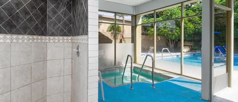 A modern indoor pool area with clear glass walls, a blue pool floor, and a shower area with dark tiles.