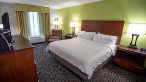 Quarto de hotel com uma cama grande, duas mesas de cabeceira, uma cadeira, uma escrivaninha, uma televisão e uma janela com cortinas.