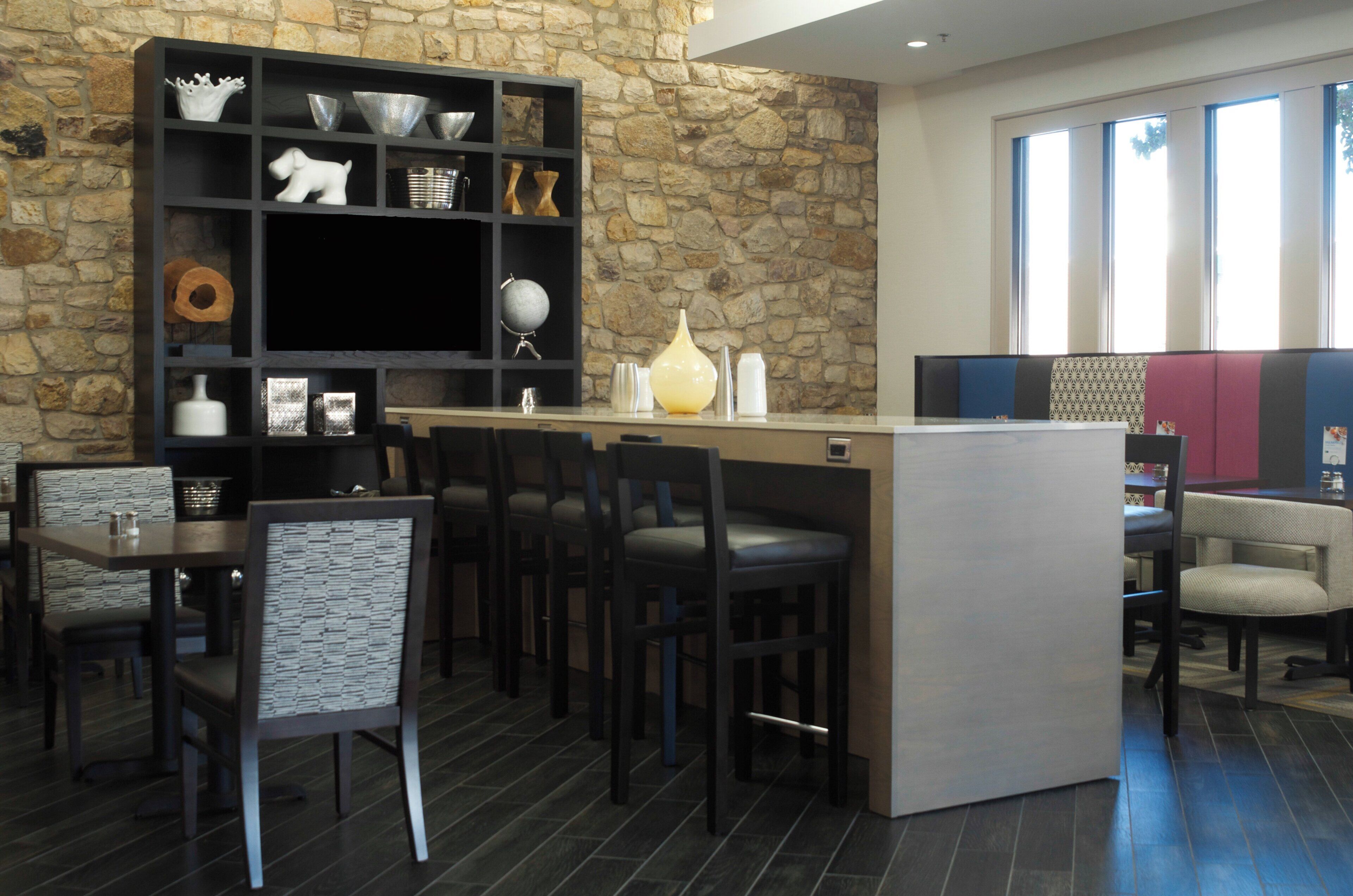 Un interior moderno con una pared de piedra, una estantería negra con varios objetos decorativos y una zona de bar con asientos.