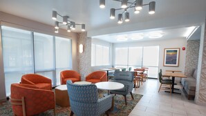 A modern hotel lobby with orange and blue patterned chairs, a round table, and large windows offering a view.