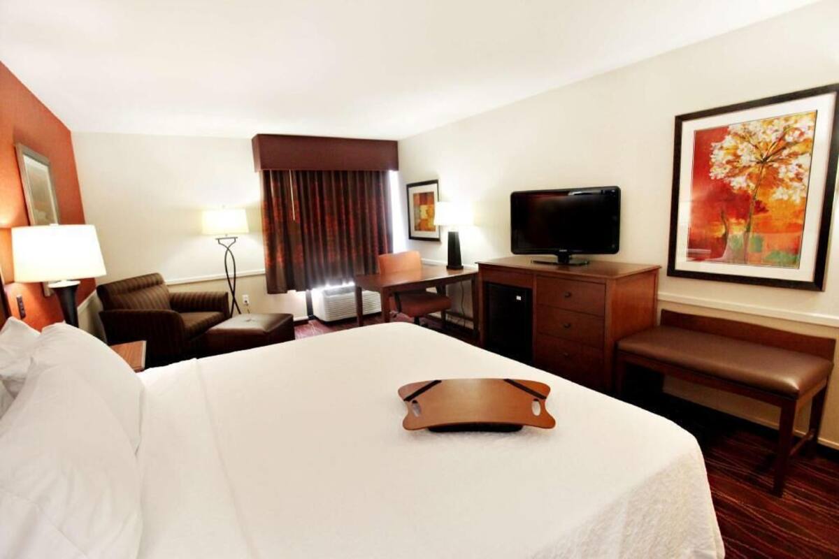 A hotel room with a bed, a desk, a television, a bench, and a painting on the wall.