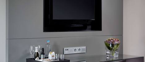 A modern hotel room with a flat-screen TV mounted on the wall, a glass desk with a tray of breakfast items, a vase with flowers, and a power outlet.