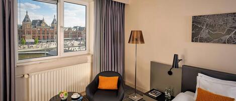 A hotel room with a bed, a chair, a small table, a lamp, and a view of a historic building through the window.