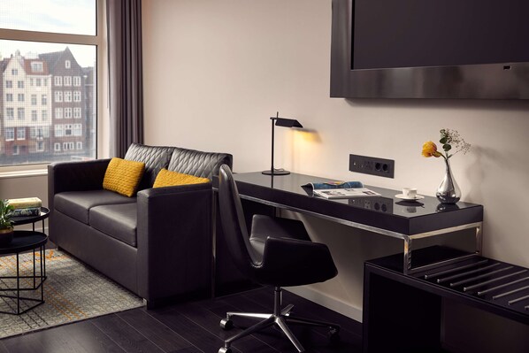 Una habitación de hotel moderna con sofá de cuero, un escritorio con lámpara, una mesita con un jarrón y vistas a edificios a través de la ventana.
